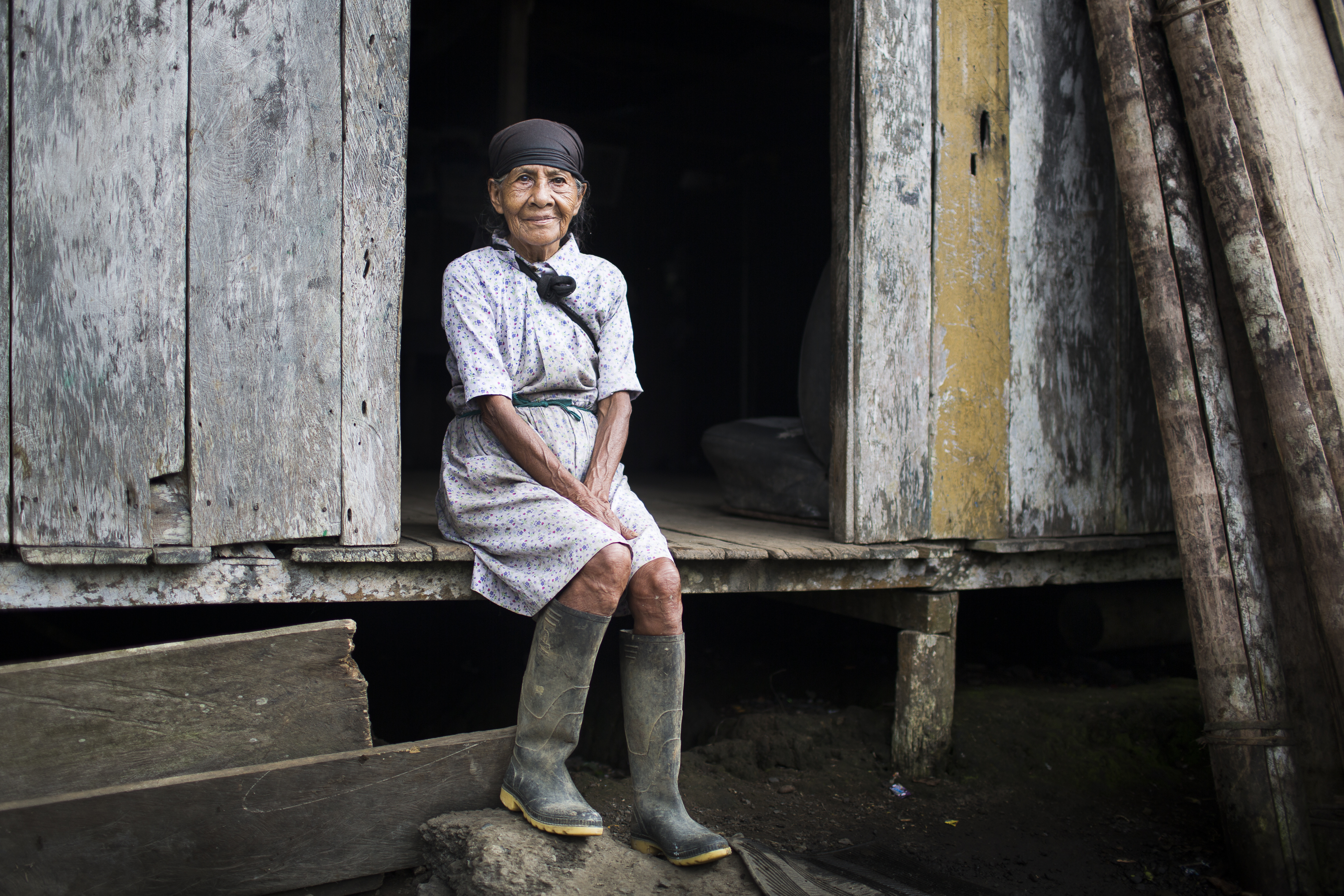 An older woman from Colombia sits at the entrance to her wooden house.
